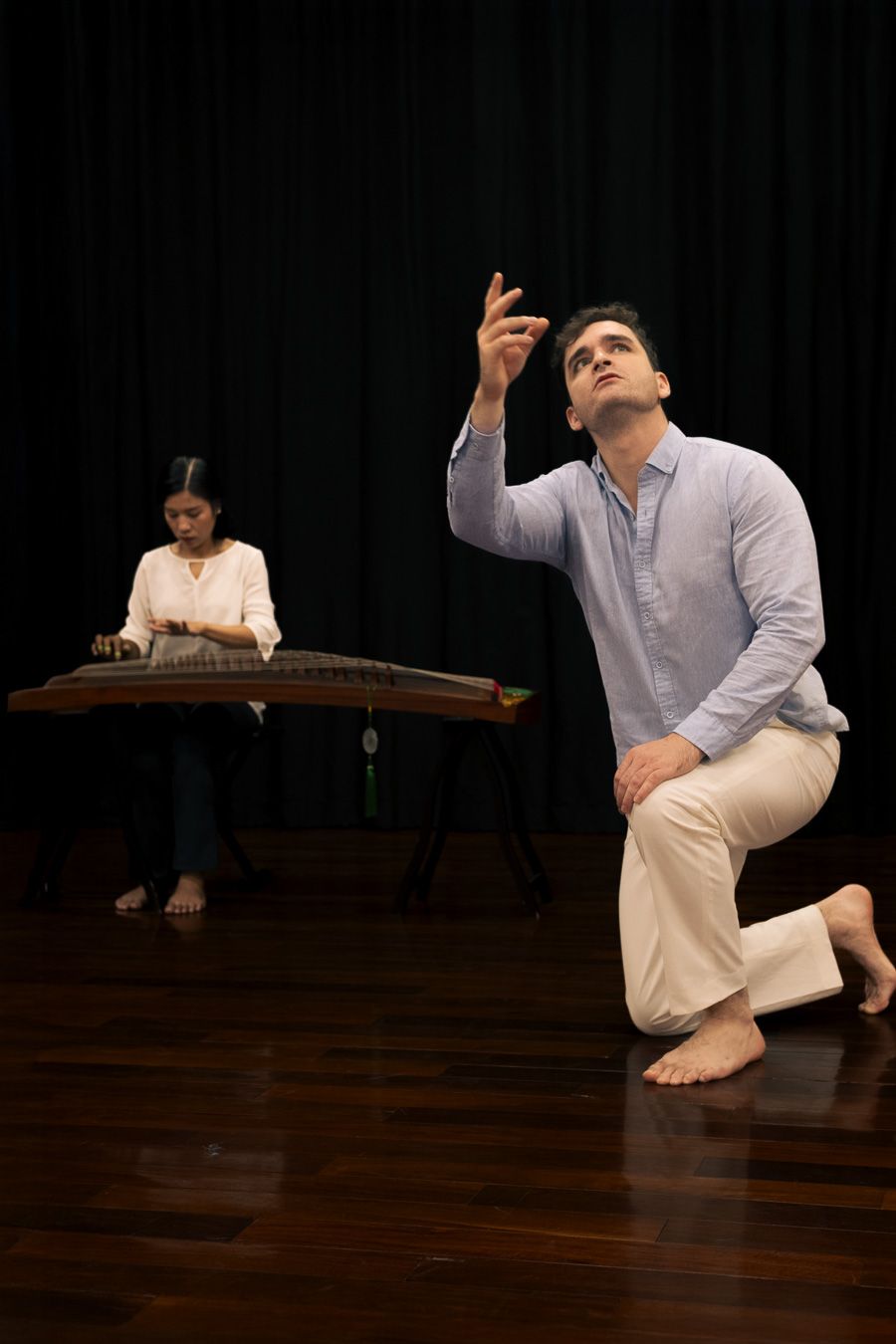Léo kneeling, looking up, hand raised, the musician playing behind
