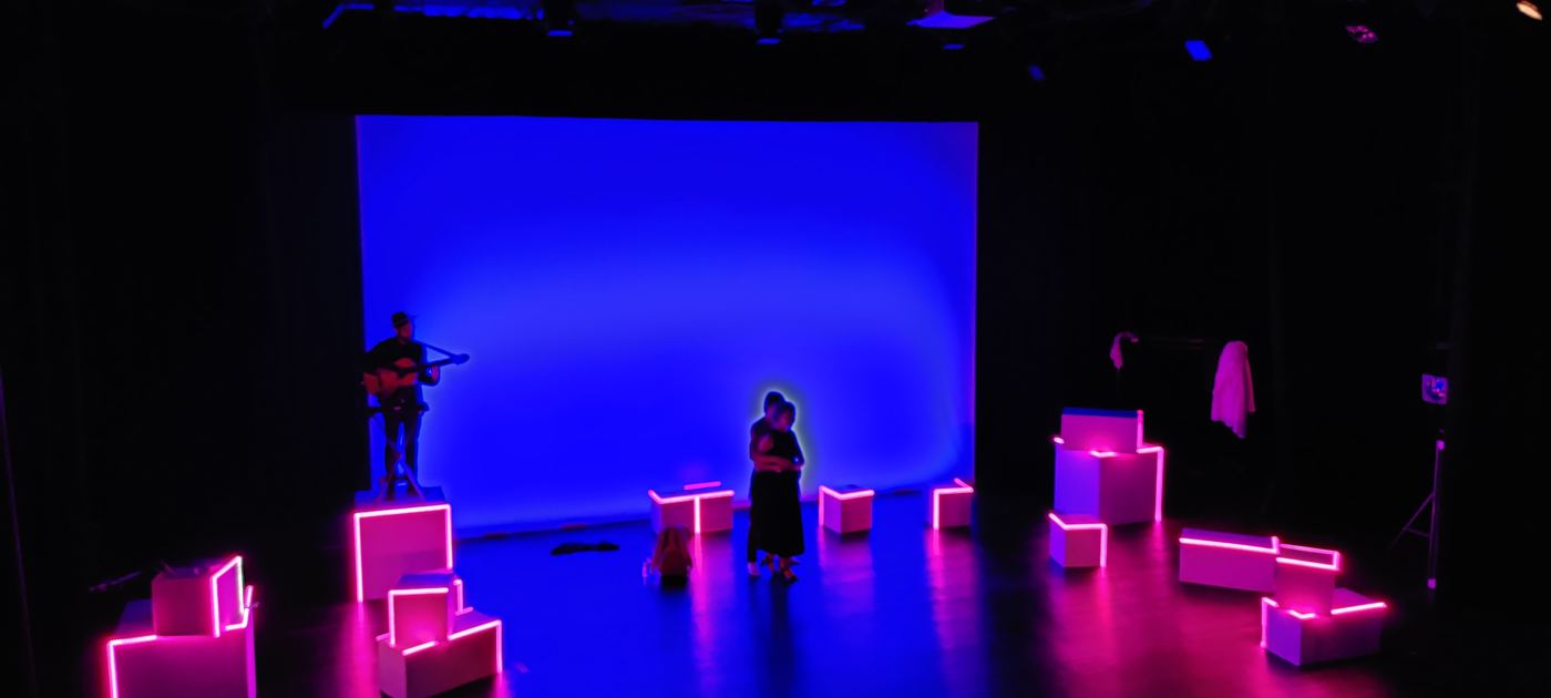 Wide shot of the stage in blue, cubes glowing magenta, a guitarist and actors