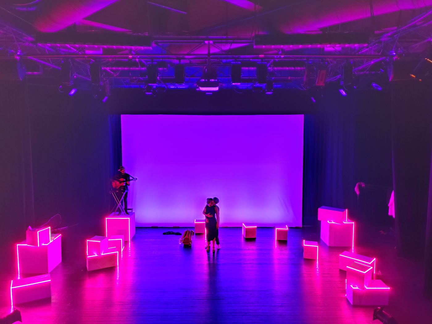 Theater stage in magenta light, luminous cubes scattered around two actors and a guitarist